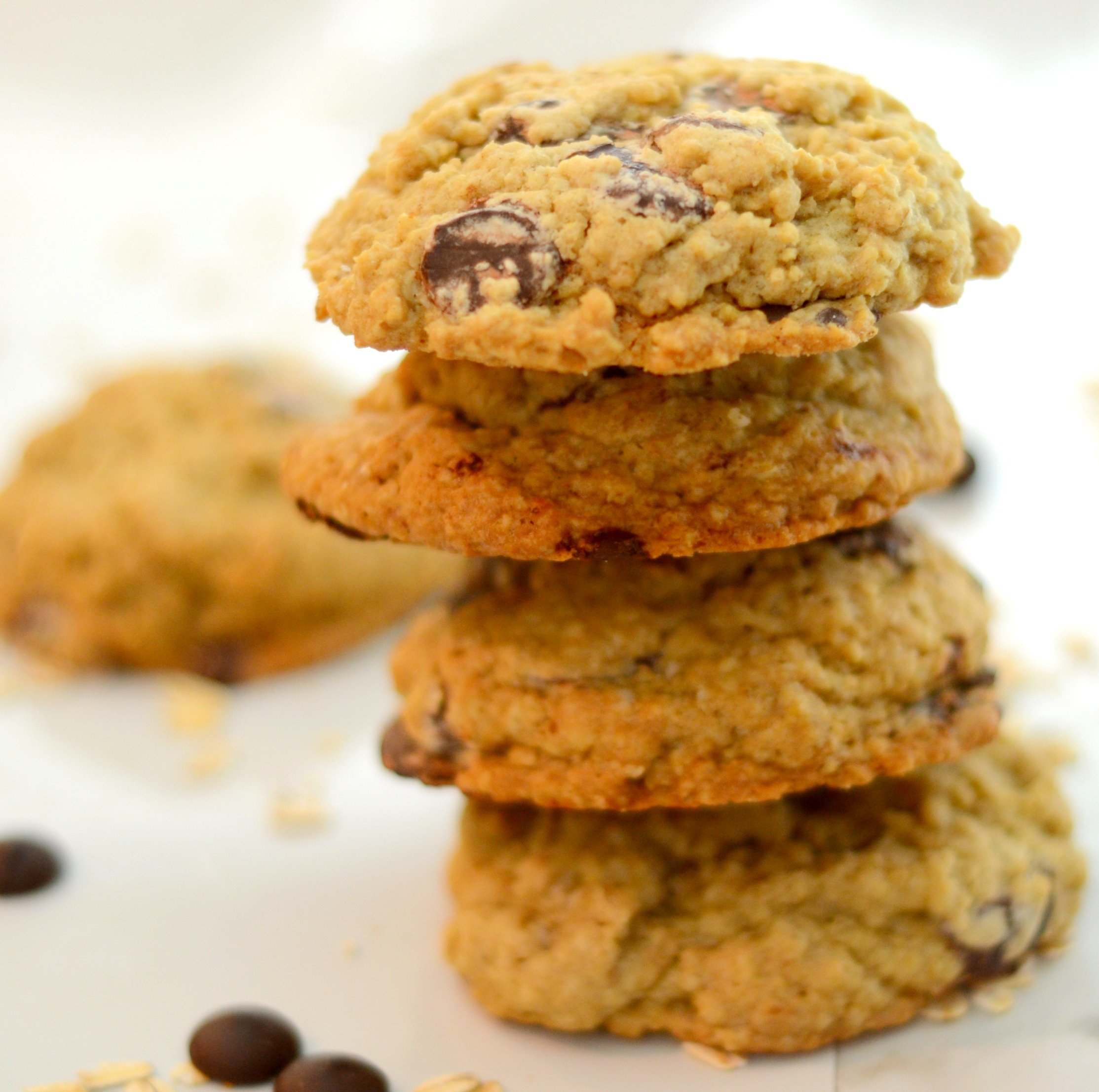 A stack of oatmeal chocolate chip cookies aganist a white background with a few chocolate chips and oats sprinkled in the foreground