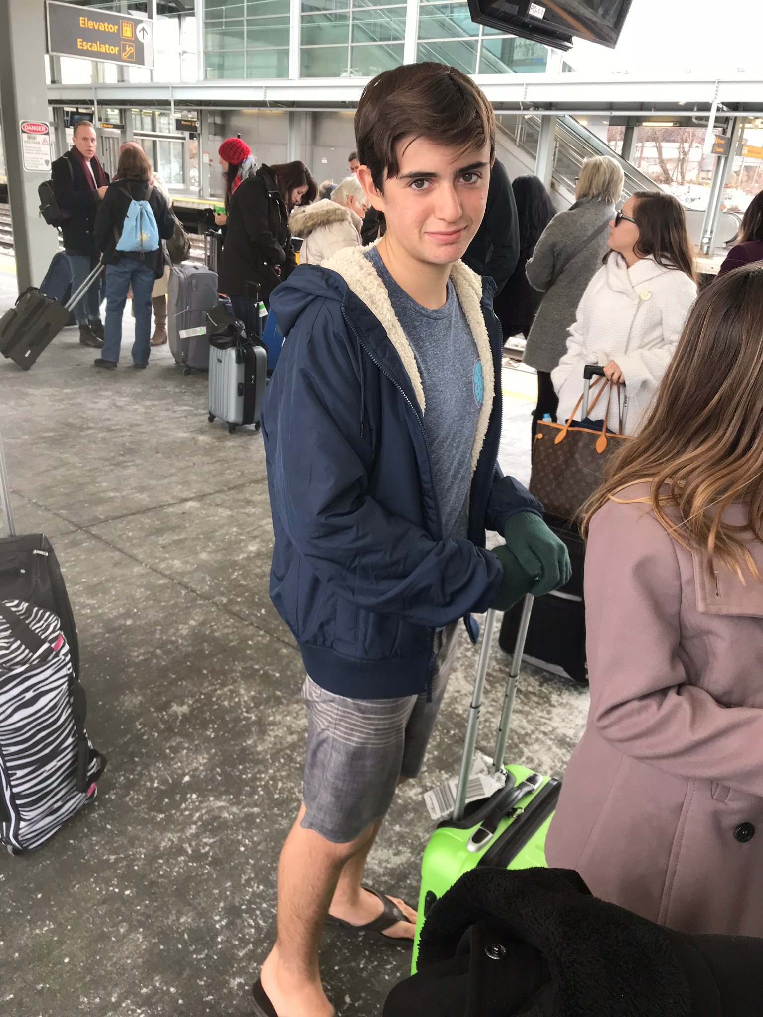 boy wearing shorts and flip flops, gloves and jacket in cold weather at a train station