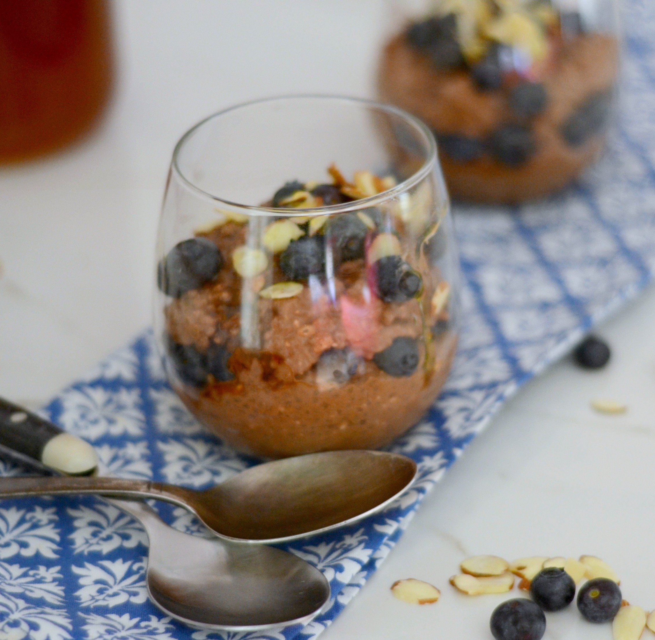 two spoons, two cups filled with chocolate chia pudding topped with blueberries and almonds, jar of honey in background