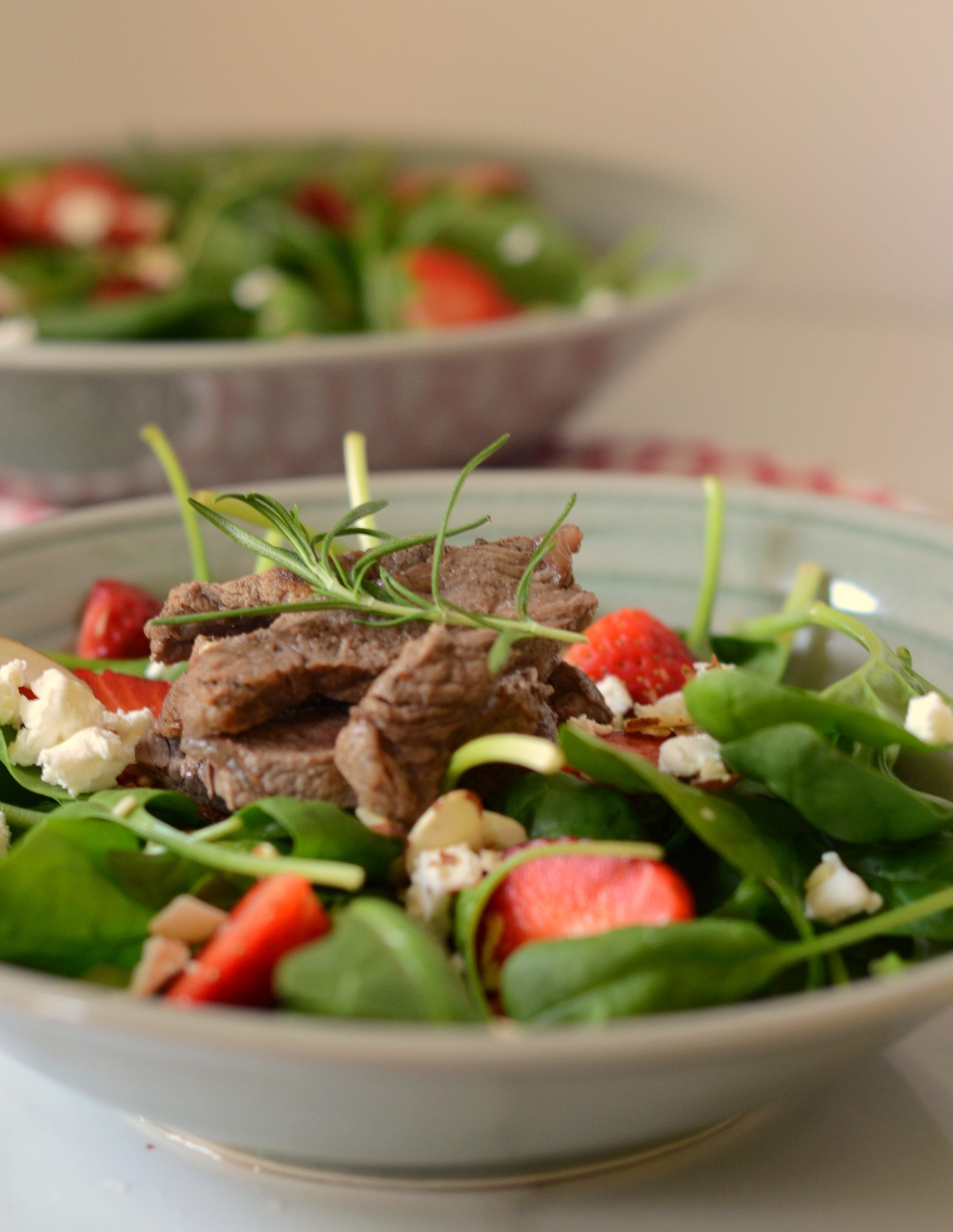 close up of a bowl of spinach and other greens topped with strawberries, steak and goat cheese