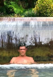 man sitting in hot spring behind waterfall